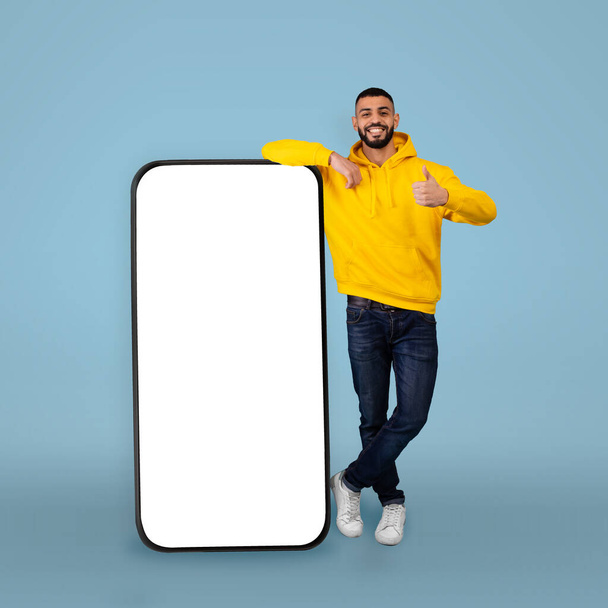 Mobile offer. Happy arab man leaning on big smartphone with blank screen and gesturing thumb up, blue background - Foto, Bild