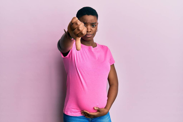 A fiatal afro-amerikai nő gyermeket vár, megérinti a terhes hasat boldogtalannak és dühösnek tűnik, elutasítást mutat, és negatív hüvelykujjal lefelé gesztussal. rossz kifejezés.  - Fotó, kép