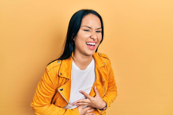 Belle femme hispanique avec piercing du nez portant une veste en cuir jaune souriant et riant fort à haute voix parce que drôle blague folle avec les mains sur le corps.  - Photo, image