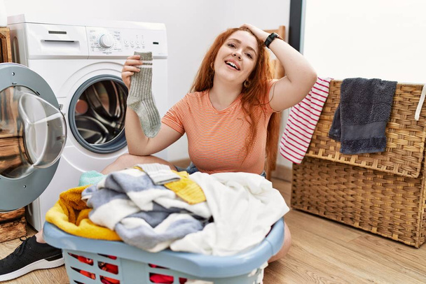 Jeune rousse femme mettre linge sale dans la machine à laver souriant confiant toucher les cheveux avec geste de la main vers le haut, posant attrayant et à la mode  - Photo, image