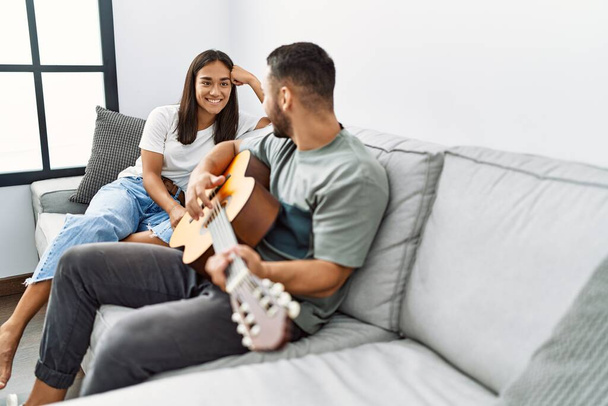 Genç latin çift evdeki kanepede oturup klasik gitar çalıyor.. - Fotoğraf, Görsel
