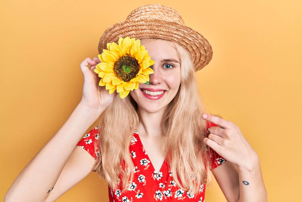Krásná běloška s blond vlasy drží žluté slunečnice přes oko usměvavý šťastný ukazující rukou a prstem  - Fotografie, Obrázek