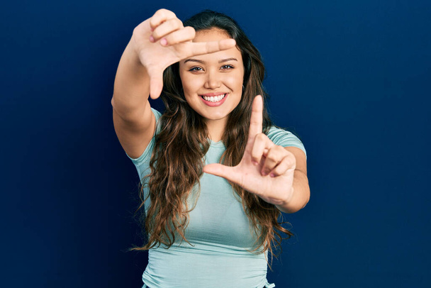 Jeune fille hispanique portant des vêtements décontractés souriant faisant cadre avec les mains et les doigts avec le visage heureux. concept de créativité et photographie.  - Photo, image