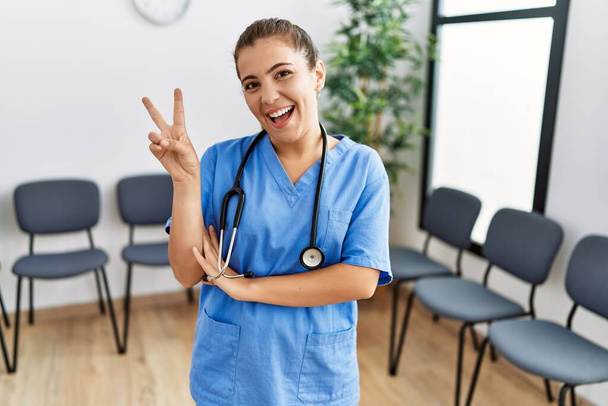 Young brunette doctor woman at waiting room smiling with happy face winking at the camera doing victory sign. number two.  - Photo, Image