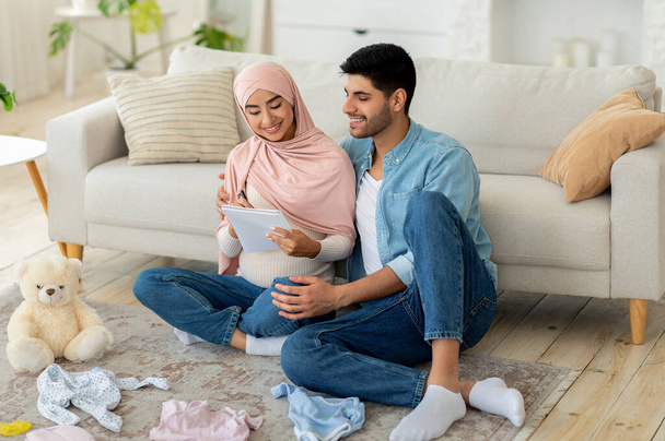 Houden van zwangere moslim paar klaar voor de bevalling, het maken van checklist tijdens het zitten met babykleding rond - Foto, afbeelding