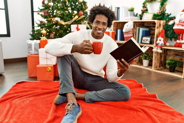 Joven afroamericano hombre leyendo libro y tomando café sentado en el suelo por árbol de Navidad en casa. - Foto, Imagen