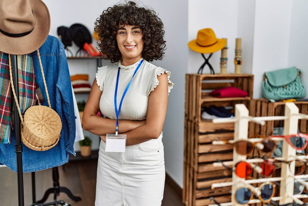 Młoda sprzedawczyni z Bliskiego Wschodu uśmiecha się szczęśliwa stojąc z rękoma skrzyżowanymi w sklepie odzieżowym. - Zdjęcie, obraz