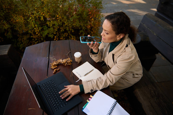 High angle view of a pretty woman, confident business lady sitting in an outdoor wooden cafeteria in front of her laptop, holding a smartphone and recording a voice message - Photo, Image