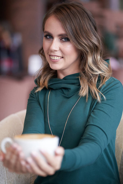 Photo d'une belle brune optimiste avec une belle coiffure, tenant une tasse de café, dans un col roulé vert, assise dans un café - Photo, image