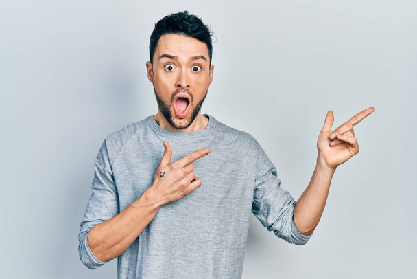 Young hispanic man pointing with fingers to the side afraid and shocked with surprise and amazed expression, fear and excited face.  - Foto, imagen