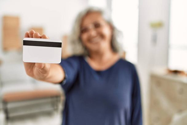 Mujer de pelo gris de mediana edad sonriendo feliz celebración de la tarjeta de crédito en el nuevo hogar. - Foto, Imagen