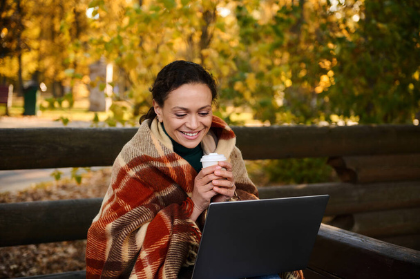Smiling middle aged woman wrapped in warm checkered blanket drinks coffee, studying , working remotely, planning projects sitting on wooden outdoor terrace of cozy cafe far from city hustle - Фото, зображення