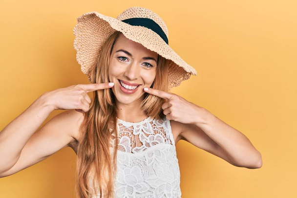 Młoda, kaukaska kobieta w letniej czapce, uśmiechnięta i wskazująca palcami zębami i ustami. Koncepcja zdrowia zębów.  - Zdjęcie, obraz