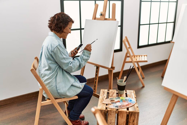 Middle age hispanic artist woman using smartphone drawing at art studio. - Фото, изображение
