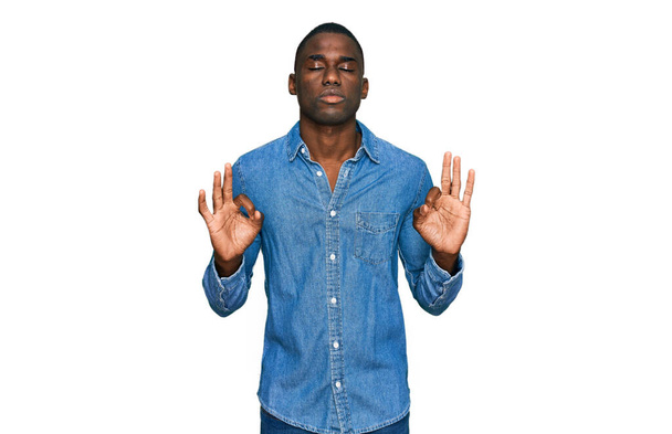 カジュアルな服を着た若いアフリカ系アメリカ人の男性は、指で瞑想ジェスチャーを行う閉じて目でリラックスして笑顔。ヨガのコンセプト.  - 写真・画像