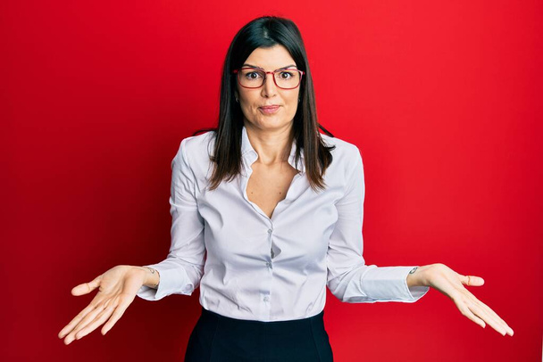 Junge hispanische Frau mit Businesshemd und Brille, ahnungslos und verwirrt mit erhobenen Armen und Händen. Zweifelhaftes Konzept.  - Foto, Bild