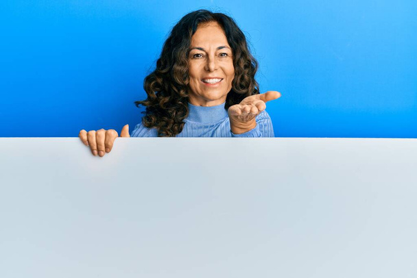 Hispanische Frau mittleren Alters hält leeres Banner in der Hand und lächelt fröhlich und bietet Handflächen an, die Hilfe und Akzeptanz geben.  - Foto, Bild