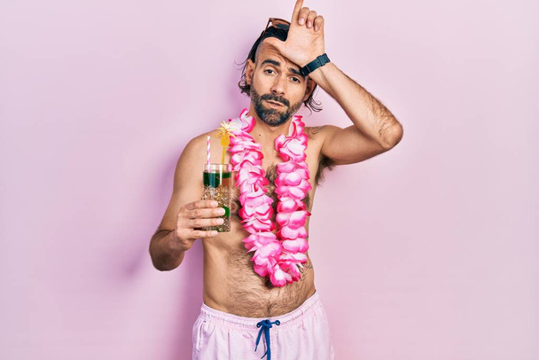 Fiatal spanyol férfi fürdőruhában és hawaii leben trópusi koktélt iszik, gúnyt űz az emberekből, ujjakkal a homlokán, lúzer gesztusokat csinál, gúnyolódik és sérteget..  - Fotó, kép