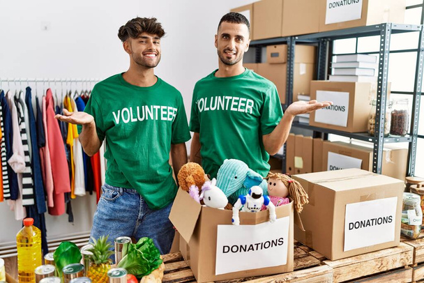 Young gay couple wearing volunteer t shirt at donations stand smiling cheerful presenting and pointing with palm of hand looking at the camera.  - Photo, Image