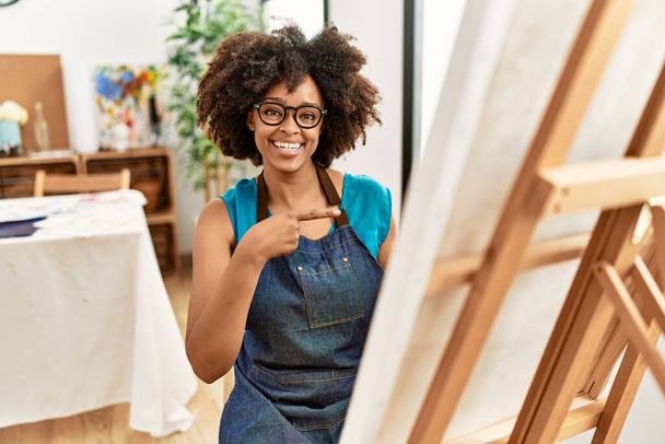 Beautiful african american woman with afro hair painting canvas at art studio smiling happy pointing with hand and finger  - Photo, Image