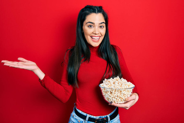 Young hispanic girl eating popcorn celebrating achievement with happy smile and winner expression with raised hand  - Fotó, kép