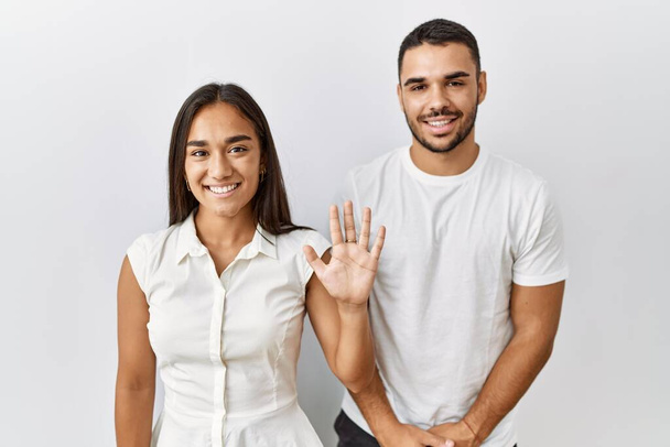 Junges gemischtrassiges Paar steht verliebt vor isoliertem Hintergrund und zeigt mit Finger Nummer fünf nach oben, während es selbstbewusst und glücklich lächelt.  - Foto, Bild