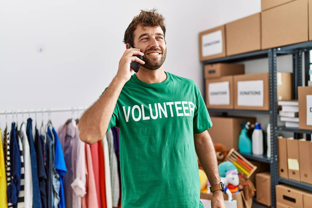 Joven voluntario hispano sonriendo feliz hablando en el teléfono inteligente en el centro de caridad. - Foto, Imagen