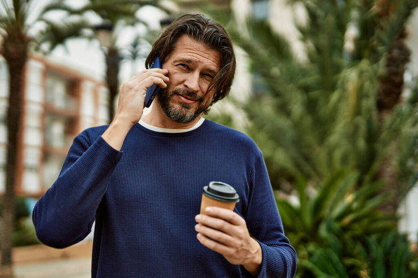 Middelbare leeftijd knappe man spreekt aan de telefoon buiten in het park - Foto, afbeelding