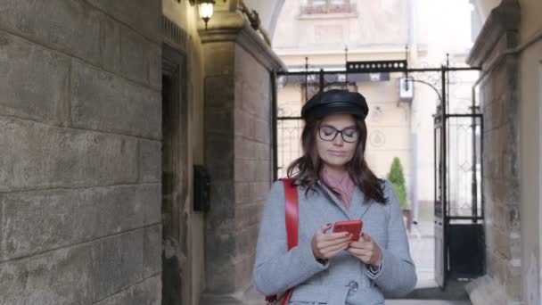  Jong mooi meisje in de herfst jurk maakt gebruik van een smartphone. Mooie vrouw loopt door de oude stad met een smartphone in haar handen - Video