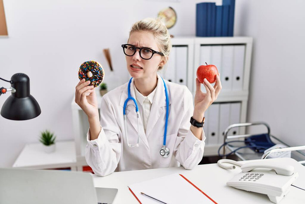 Junge Ärztin mit rotem Apfel und Donut in der Klinik ahnungslos und verwirrt. Zweifelhaftes Konzept.  - Foto, Bild