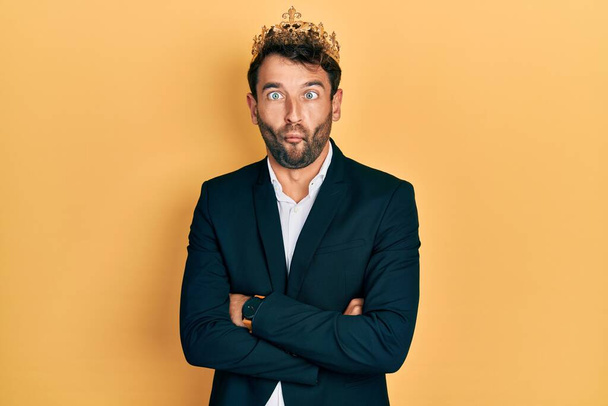 Handsome man with beard wearing business suit with arms crossed gesture and king crown making fish face with mouth and squinting eyes, crazy and comical.  - Foto, imagen