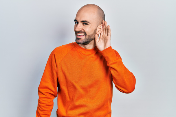 Young bald man wearing casual clothes smiling with hand over ear listening and hearing to rumor or gossip. deafness concept.  - Photo, Image