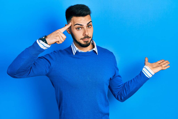 Young hispanic man with beard wearing casual blue sweater confused and annoyed with open palm showing copy space and pointing finger to forehead. think about it.  - Photo, Image