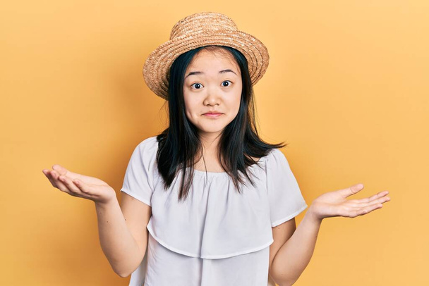 Giovane ragazza cinese che indossa cappello estivo espressione indiscreta e confusa con le braccia e le mani alzate. concetto di dubbio.  - Foto, immagini