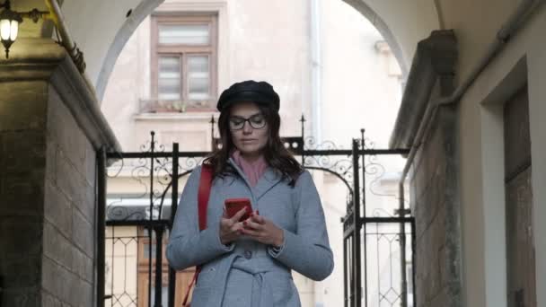Ligubella ragazza in abito autunnale utilizza uno smartphone. Bella donna cammina attraverso la città vecchia con uno smartphone in mano - Filmati, video
