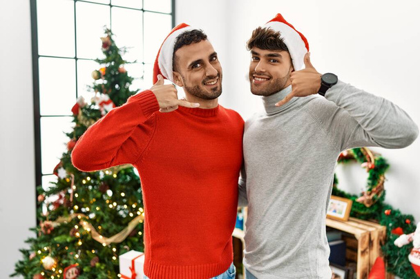 Young gay couple standing by christmas tree wearing hat smiling doing phone gesture with hand and fingers like talking on the telephone. communicating concepts.  - Photo, Image
