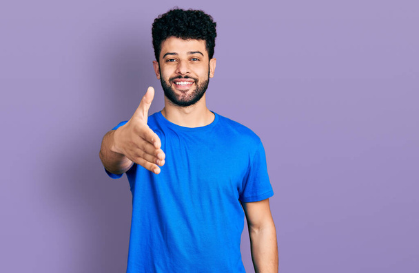 Hombre árabe joven con barba con camiseta azul casual sonriendo amigable ofreciendo apretón de manos como saludo y bienvenida. negocios exitosos.  - Foto, imagen