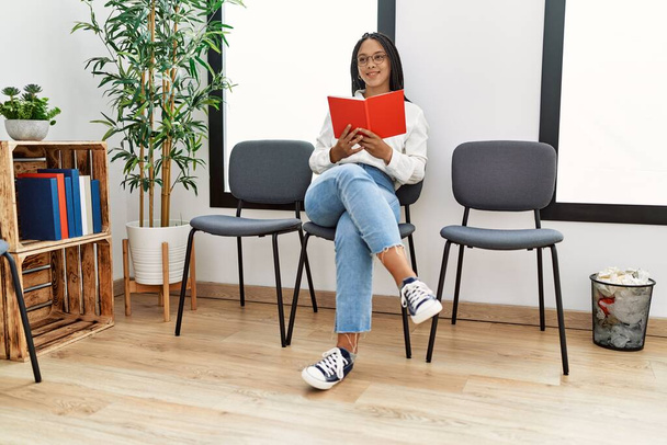 Νεαρή Αφροαμερικανή γυναίκα χαμογελά αυτοπεποίθηση διαβάζοντας βιβλίο στην αίθουσα αναμονής - Φωτογραφία, εικόνα