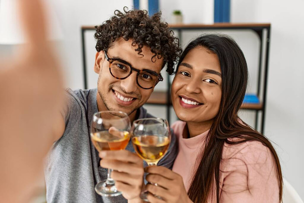 Young latin couple make selfie by the camera toasting with wine sitting on the table at home. - Fotoğraf, Görsel