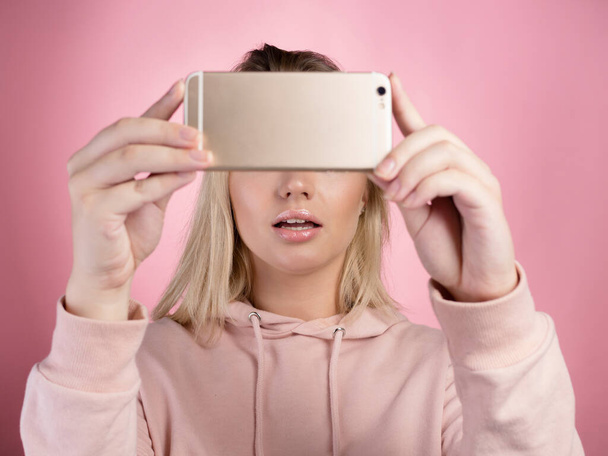 attractive young woman in a pink hoodie uses a smartphone for video communication or photos. - Fotoğraf, Görsel