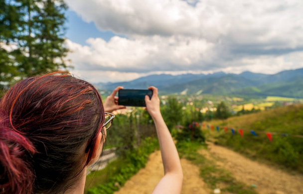 Back view of woman holding smartphone in both hands during hike, taking photos for travel blog, looking for new route online, searching for connection in mountain tour. Copyspace for advertisement - Photo, Image