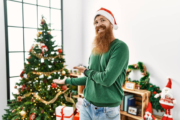 Redhead man with long beard wearing christmas hat by christmas tree inviting to enter smiling natural with open hand  - Zdjęcie, obraz