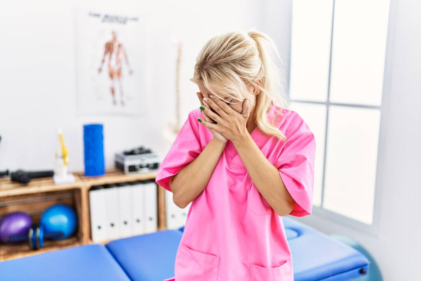 Jonge blanke vrouw die werkt in een pijnkliniek met een droevige uitdrukking en gezicht bedekt met handen terwijl ze huilt. depressie concept.  - Foto, afbeelding