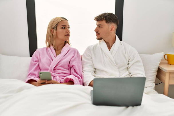 若い白人のカップルは、自宅でベッドの上に座ってラップトップとスマートフォンを使用して真剣な表情で. - 写真・画像