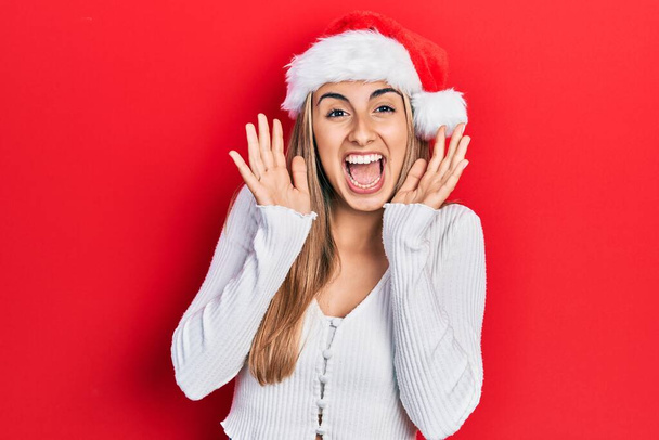 Piękna latynoska kobieta w świątecznym kapeluszu świętująca szaleństwo i zdumiona sukcesem z podniesionymi ramionami i otwartymi oczami krzyczącymi podekscytowana. koncepcja zwycięzcy  - Zdjęcie, obraz