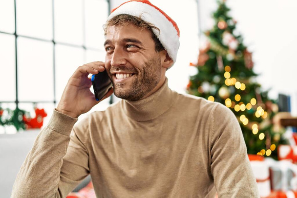 Junger hispanischer Mann spricht auf dem Smartphone und sitzt zu Hause am Weihnachtsbaum - Foto, Bild