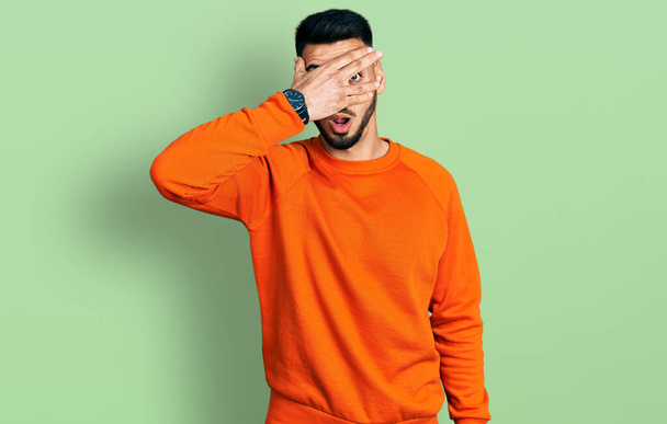 Νεαρός Ισπανός με γενειάδα που φοράει πορτοκαλί πουλόβερ κρυφοκοιτάζει στο πρόσωπο και τα μάτια με το χέρι, κοιτάζοντας μέσα από τα δάχτυλα με αμηχανία.  - Φωτογραφία, εικόνα