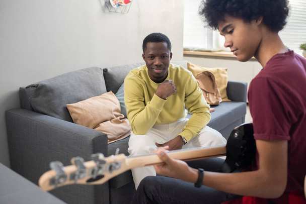 Щасливий чорношкірий в повсякденному одязі дивиться на молодого хлопця, який грає на гітарі
 - Фото, зображення