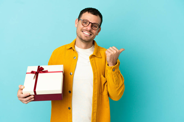 Brazilian man holding a gift over isolated blue background pointing to the side to present a product - Fotografie, Obrázek
