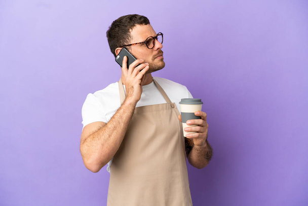 Brazilian restaurant waiter over isolated purple background holding coffee to take away and a mobile - Photo, Image